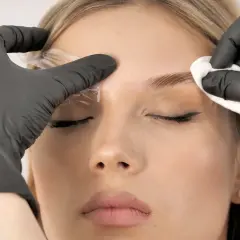 Profesjonalna laminacja brwi. Stylistka w czarnych rękawiczkach aplikuje produkt, by nadać brwiom idealny kształt.