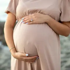 Brzuszek ciężarnej kobiety w beżowej sukience, dłonie delikatnie go obejmują. Warto pamiętać o bezpieczeństwie, np. unikając retinolu w ciąży.