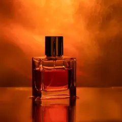 Szklana buteleczka perfum z bursztynowym płynem, otoczona ciepłym, pomarańczowym światłem. Ten zapach to obietnica luksusu.