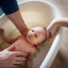 Delikatna kąpiel noworodka w wanience. Rodzice z troską trzymają dziecko, które wygląda na spokojne. Woda jest czysta, a w tle widać drewnianą podłogę. Może dodano krochmal do kąpieli dla miękkości skóry?