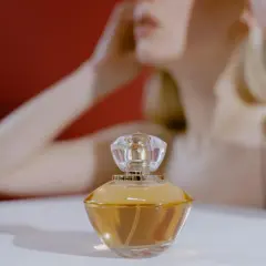 Elegancki flakonik z bursztynowym **attarem** na białym tle, w tle rozmyta postać kobiety.