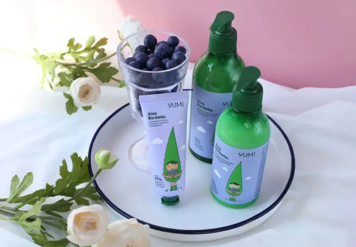 Krem do rąk i balsam do ciała YUMI Aloe Borówka, z uroczą grafiką, obok szklanki jagód i kwiatów.