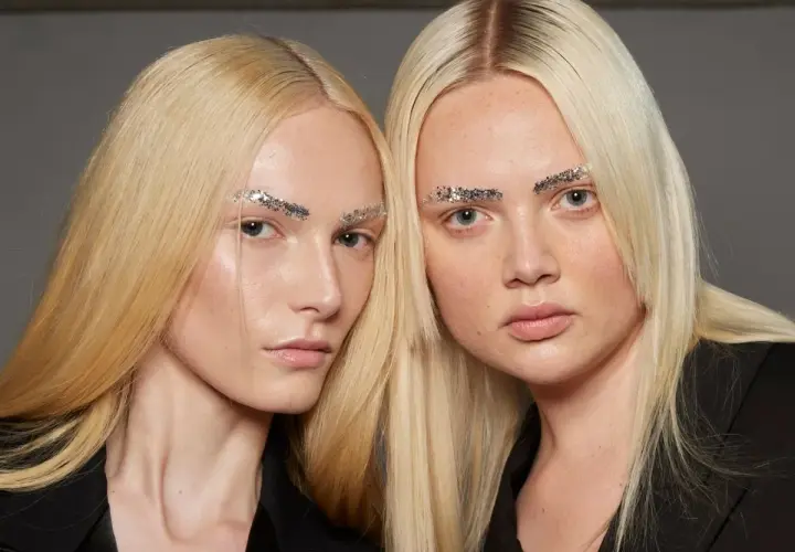 Dwie modelki o żółtym blond włosach i błyszczących brwiach z brokatem.