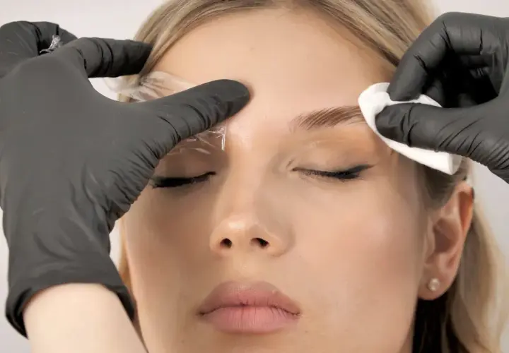 Profesjonalna laminacja brwi. Stylistka w czarnych rękawiczkach aplikuje produkt, by nadać brwiom idealny kształt.