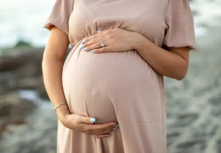 Brzuszek ciężarnej kobiety w beżowej sukience, dłonie delikatnie go obejmują. Warto pamiętać o bezpieczeństwie, np. unikając retinolu w ciąży.