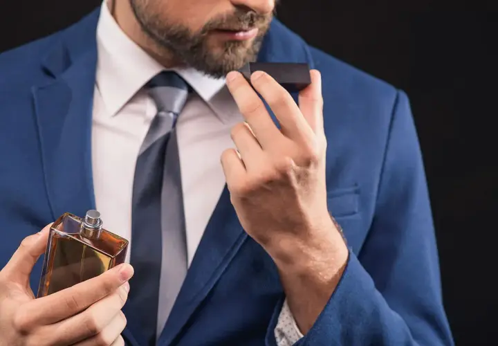 Elegancki mężczyzna w niebieskiej marynarce wącha perfumy. Zapach, niczym karnozyna, dodaje mu pewności siebie.
