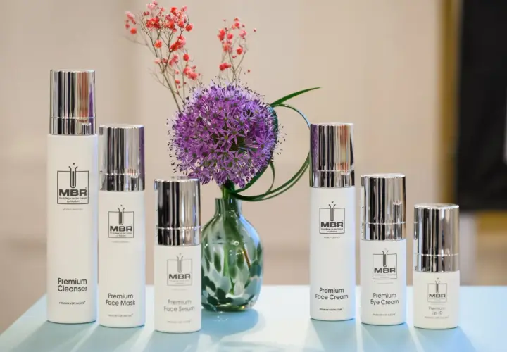 Linia kosmetyków MBR: Premium Cleanser, Face Mask, Face Serum, Face Cream, Eye Cream i Lip-ID, obok fioletowego czosnku i drobnych różowych kwiatów.