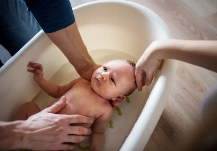 Delikatna kąpiel noworodka w wanience. Rodzice z troską trzymają dziecko, które wygląda na spokojne. Woda jest czysta, a w tle widać drewnianą podłogę. Może dodano krochmal do kąpieli dla miękkości skóry?