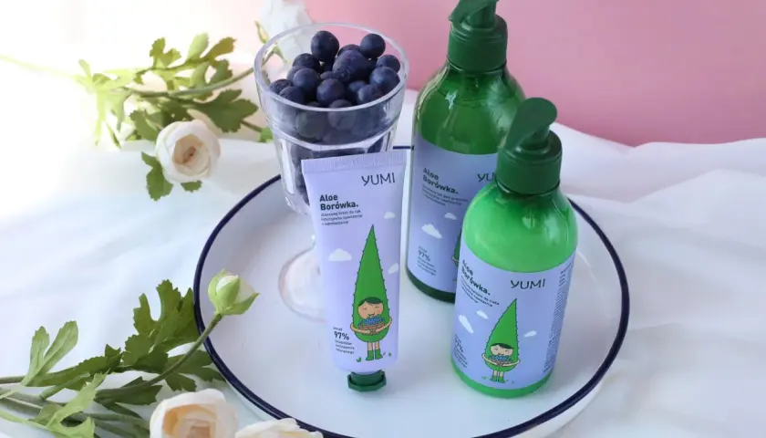 Krem do rąk i balsam do ciała YUMI Aloe Borówka, z uroczą grafiką, obok szklanki jagód i kwiatów.