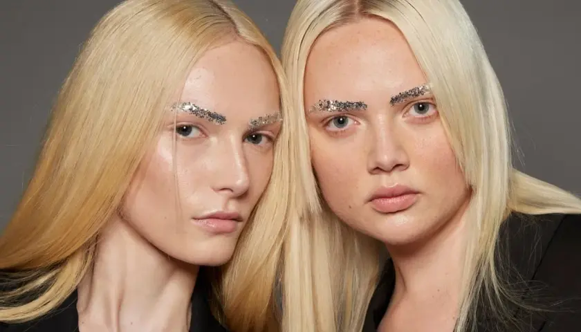 Dwie modelki o żółtym blond włosach i błyszczących brwiach z brokatem.