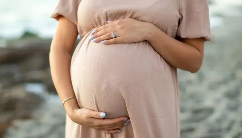 Brzuszek ciężarnej kobiety w beżowej sukience, dłonie delikatnie go obejmują. Warto pamiętać o bezpieczeństwie, np. unikając retinolu w ciąży.