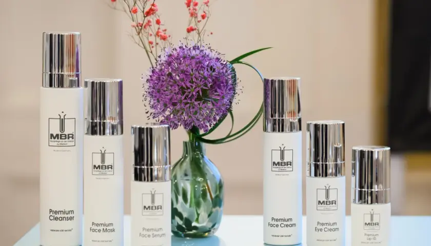 Linia kosmetyków MBR: Premium Cleanser, Face Mask, Face Serum, Face Cream, Eye Cream i Lip-ID, obok fioletowego czosnku i drobnych różowych kwiatów.