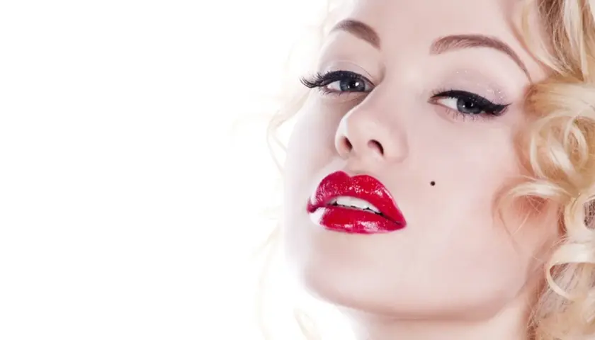Kobieta z burzą blond loków i charakterystycznym pieprzykiem, usta w stylu Marilyn Monroe, błyszczące czerwone.