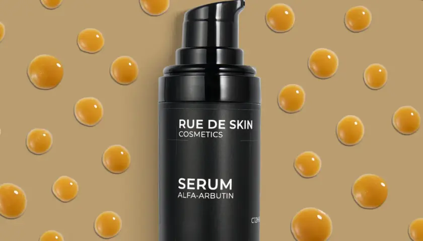 Czarny flakonik serum z alfa arbutyną, otoczony kroplami o bursztynowym kolorze, na beżowym tle.