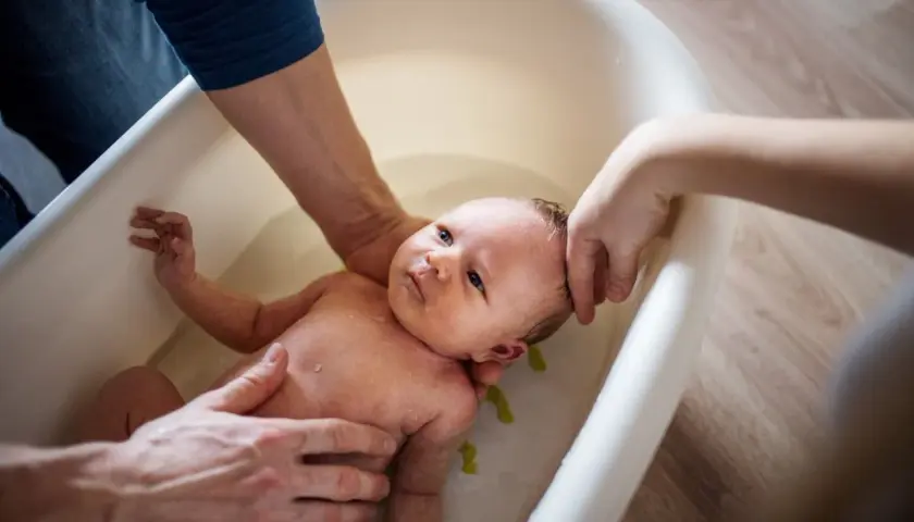 Delikatna kąpiel noworodka w wanience. Rodzice z troską trzymają dziecko, które wygląda na spokojne. Woda jest czysta, a w tle widać drewnianą podłogę. Może dodano krochmal do kąpieli dla miękkości skóry?