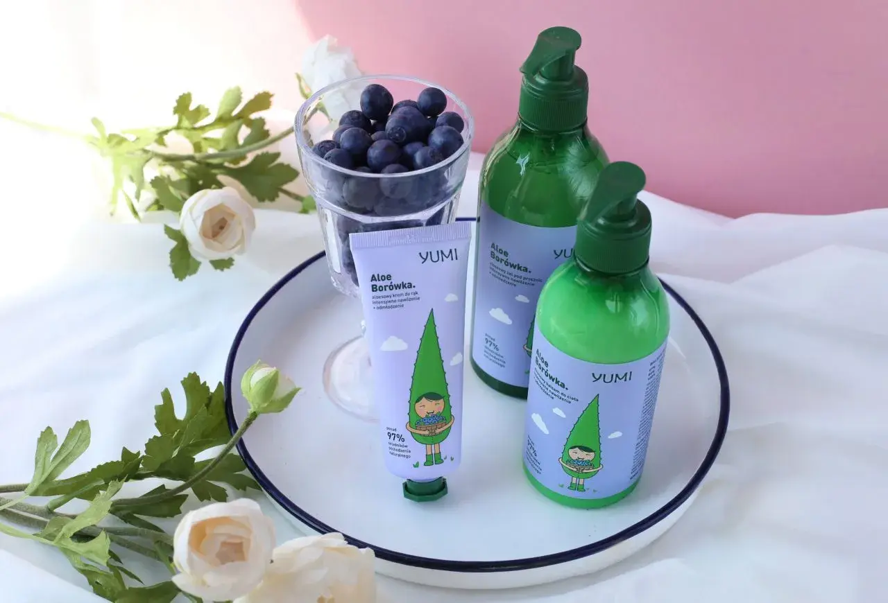 Krem do rąk i balsam do ciała YUMI Aloe Borówka, z uroczą grafiką, obok szklanki jagód i kwiatów.