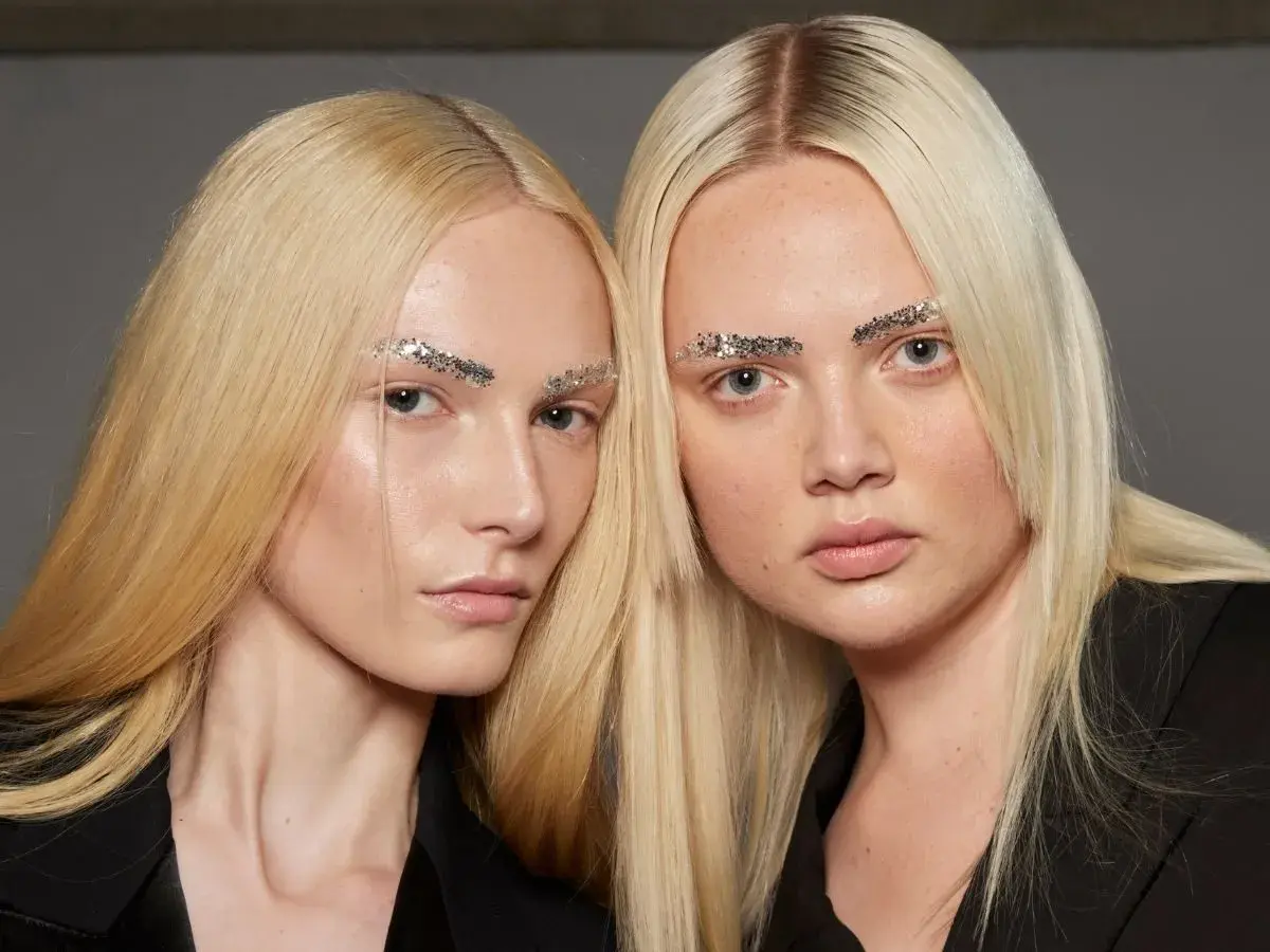 Dwie modelki o żółtym blond włosach i błyszczących brwiach z brokatem.