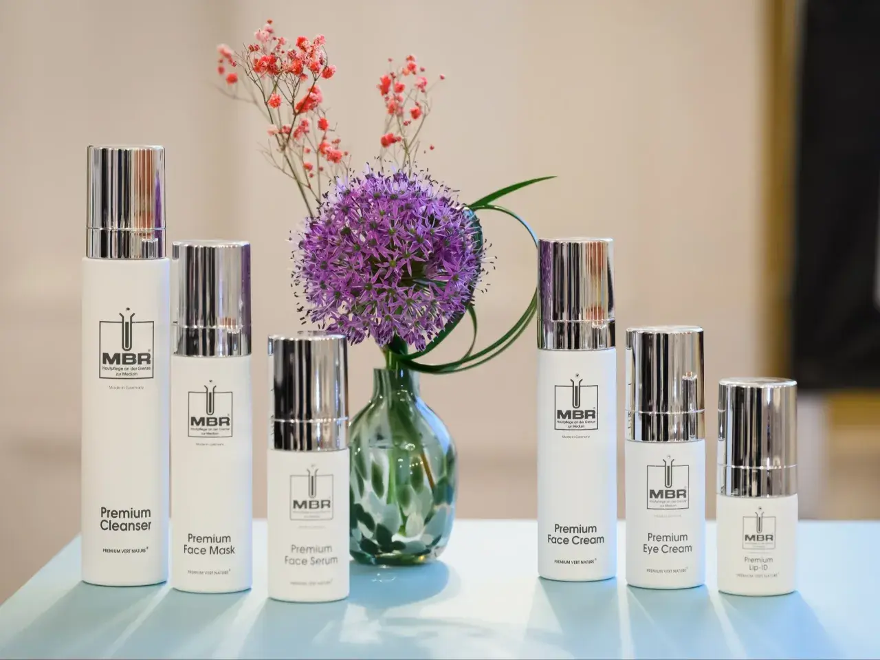 Linia kosmetyków MBR: Premium Cleanser, Face Mask, Face Serum, Face Cream, Eye Cream i Lip-ID, obok fioletowego czosnku i drobnych różowych kwiatów.