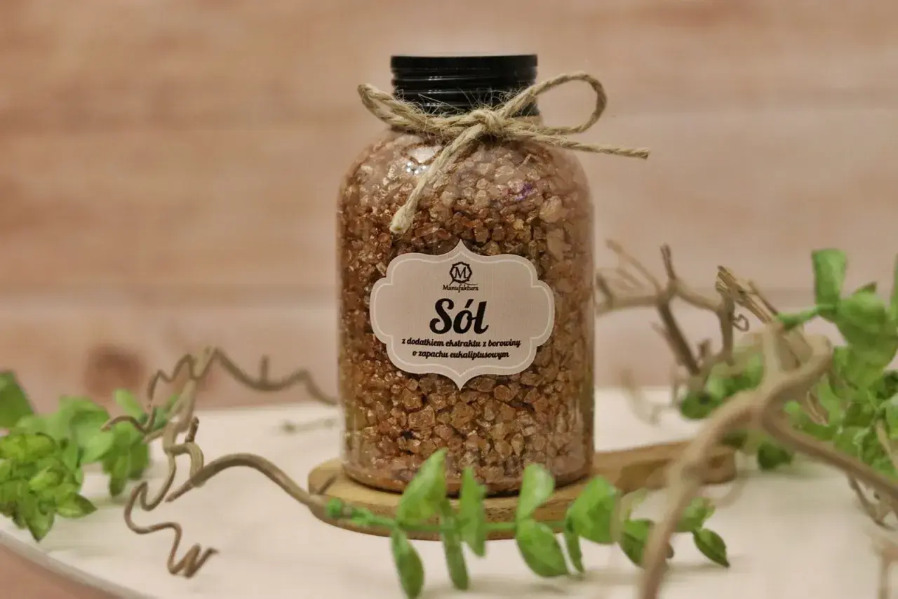 Sól borowinowa w słoiku, ozdobiona sznurkiem i etykietą. W tle drewniana deska i zielone gałązki.