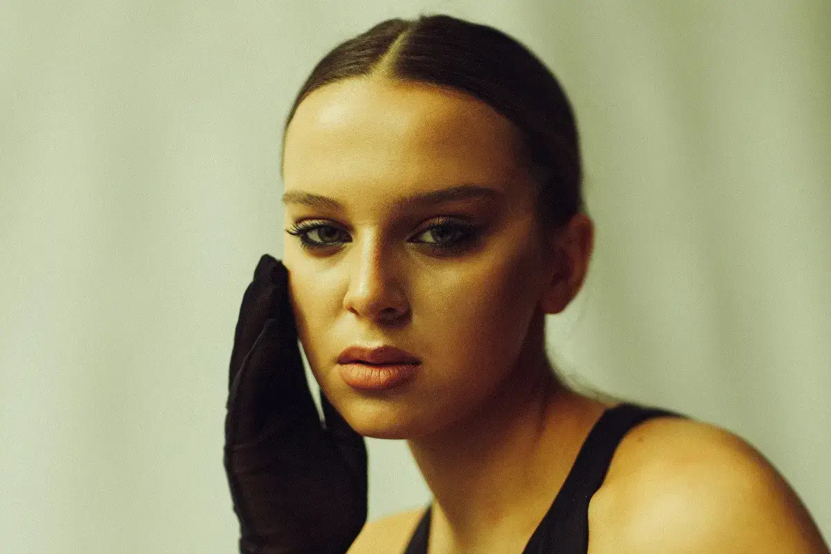 Młoda it girl w czarnej rękawiczce dotyka policzka, zmysłowy makijaż oczu i ust.