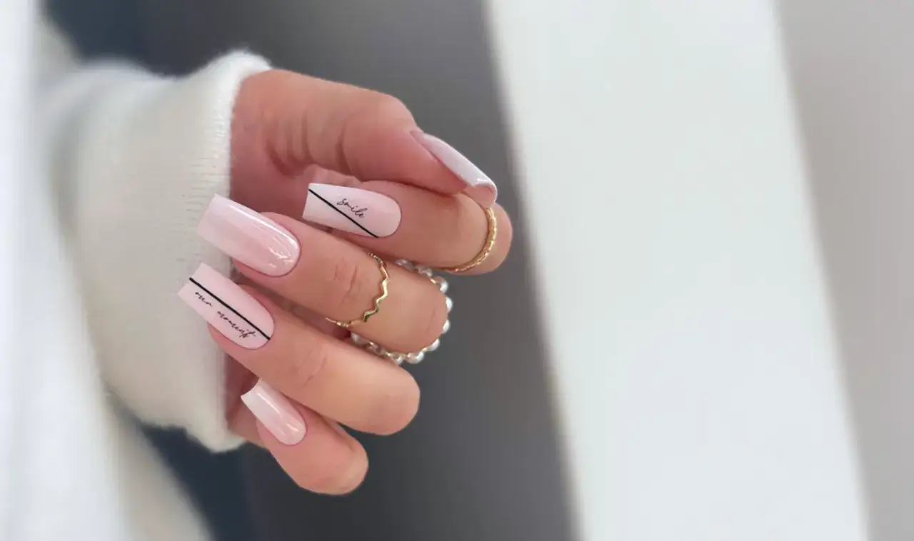 Dłoń z długimi, kwadratowymi paznokciami w odcieniach różu i bieli. Na niektórych paznokciach widnieją czarne napisy i linie. Te milky nails zdobią złote pierścionki.