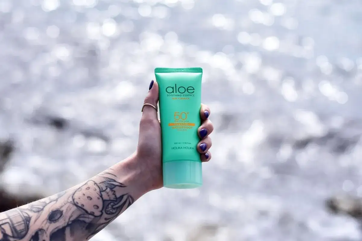 Ręka z tatuażem trzyma tubkę kremu z filtrem SPF 50+ Aloe Soothing Essence. Idealny wybór, jaki SPF do twarzy na wakacje!